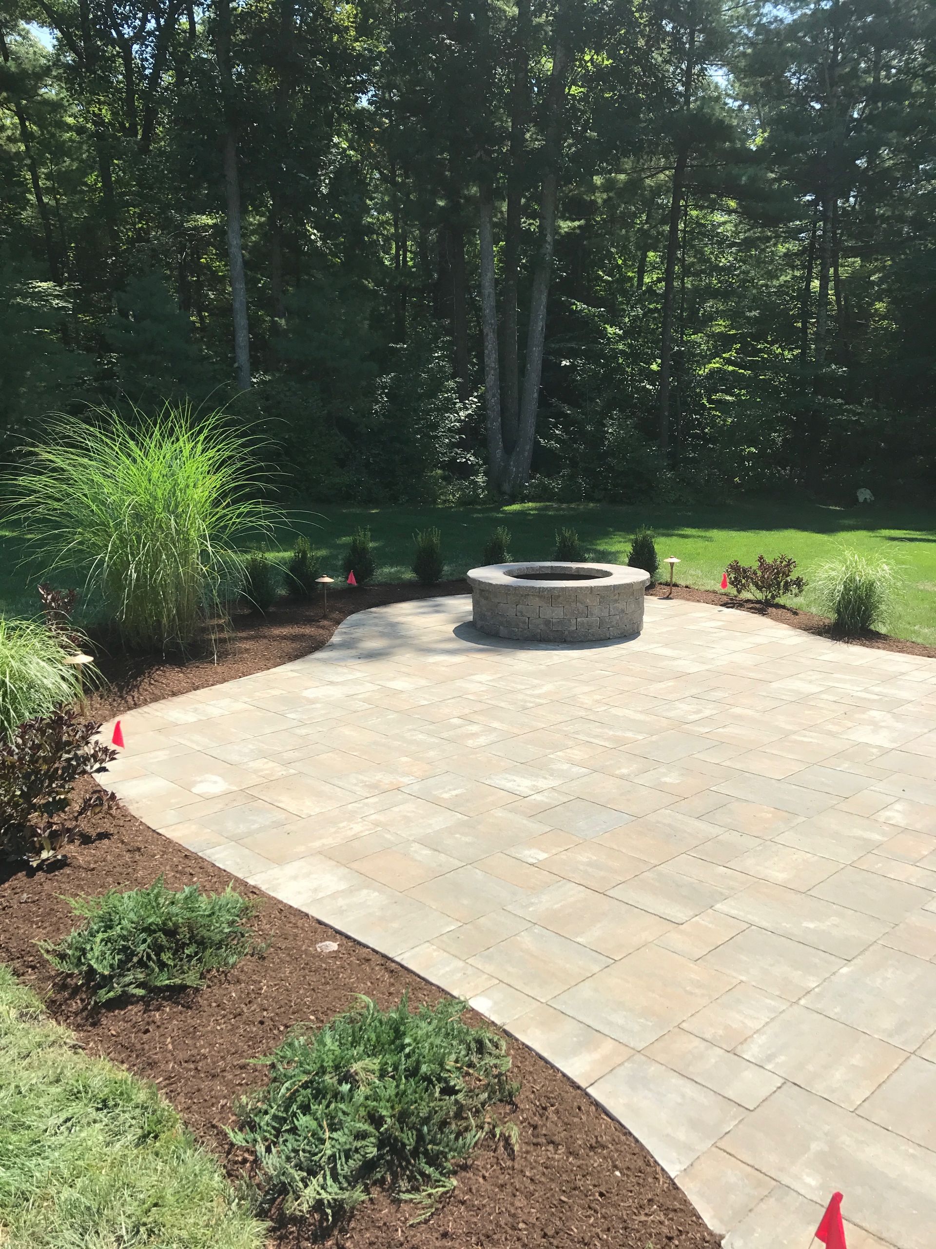 Patio with stone fire pit, bordered by shrubs, beside a wooded backyard lawn