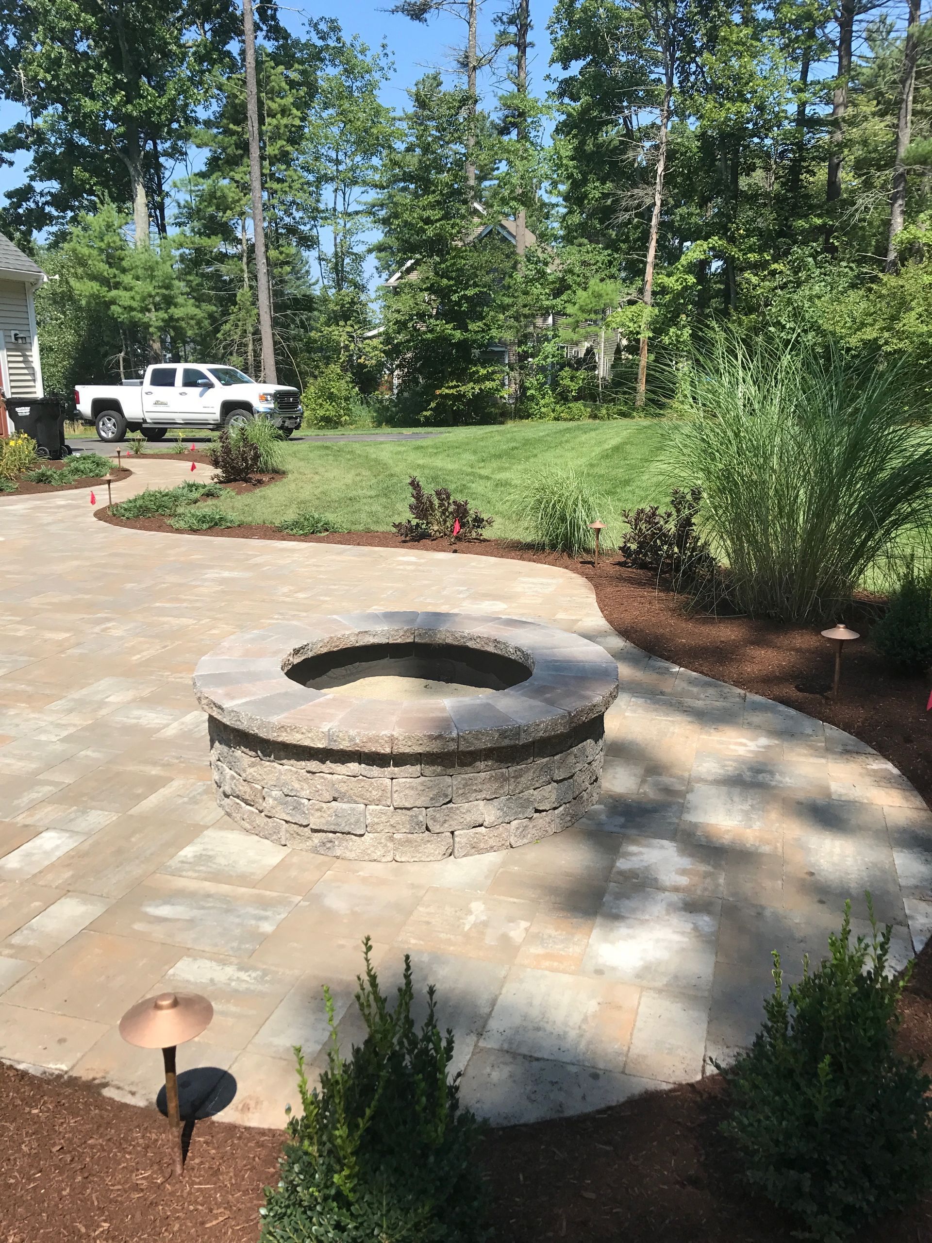 Stone fire pit on a paved patio in a sunny backyard with trees and a parked white truck in the background