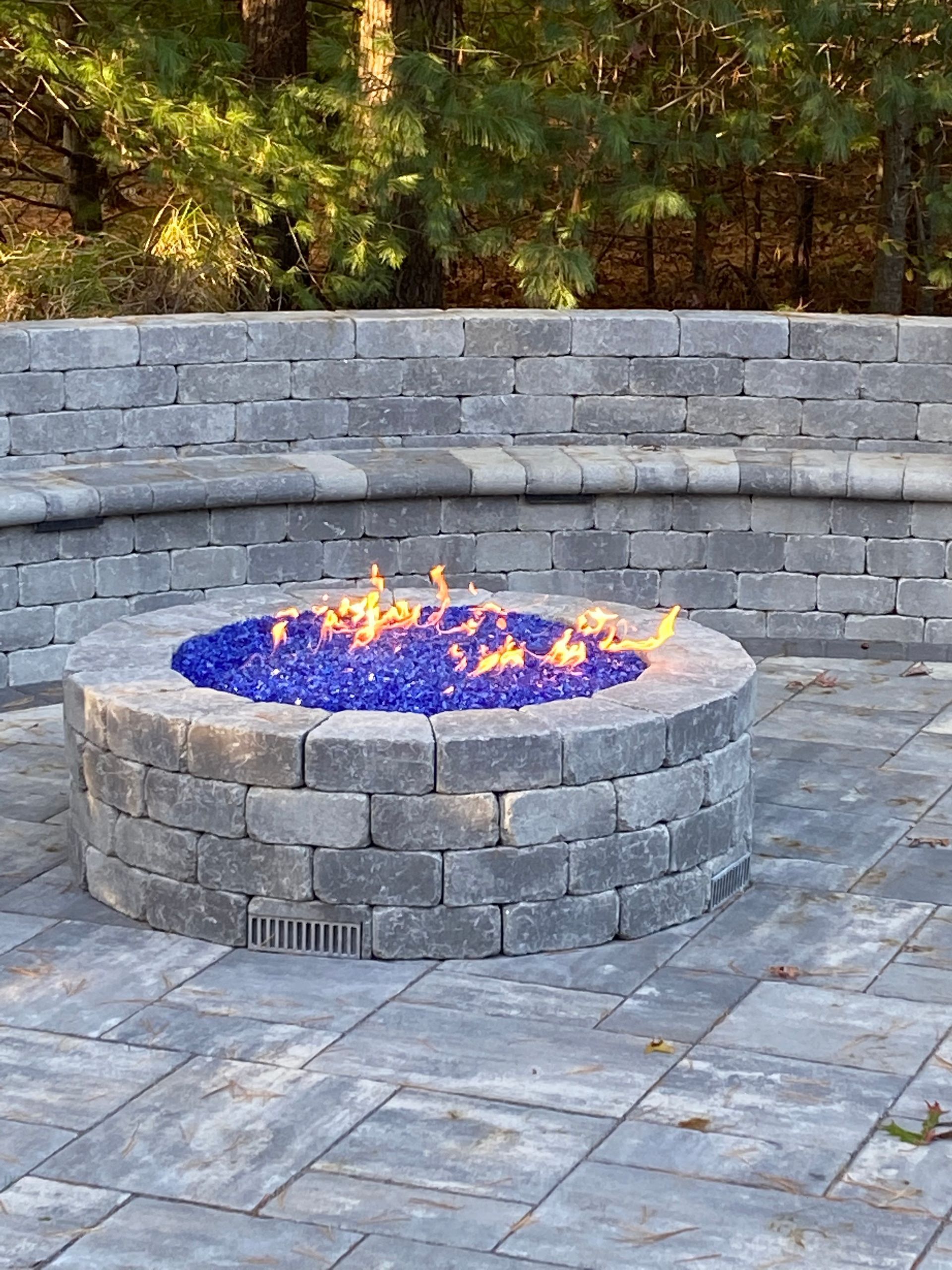 Stone fire pit with blue flames in a circular patio, backed by a curved stone wall and trees.