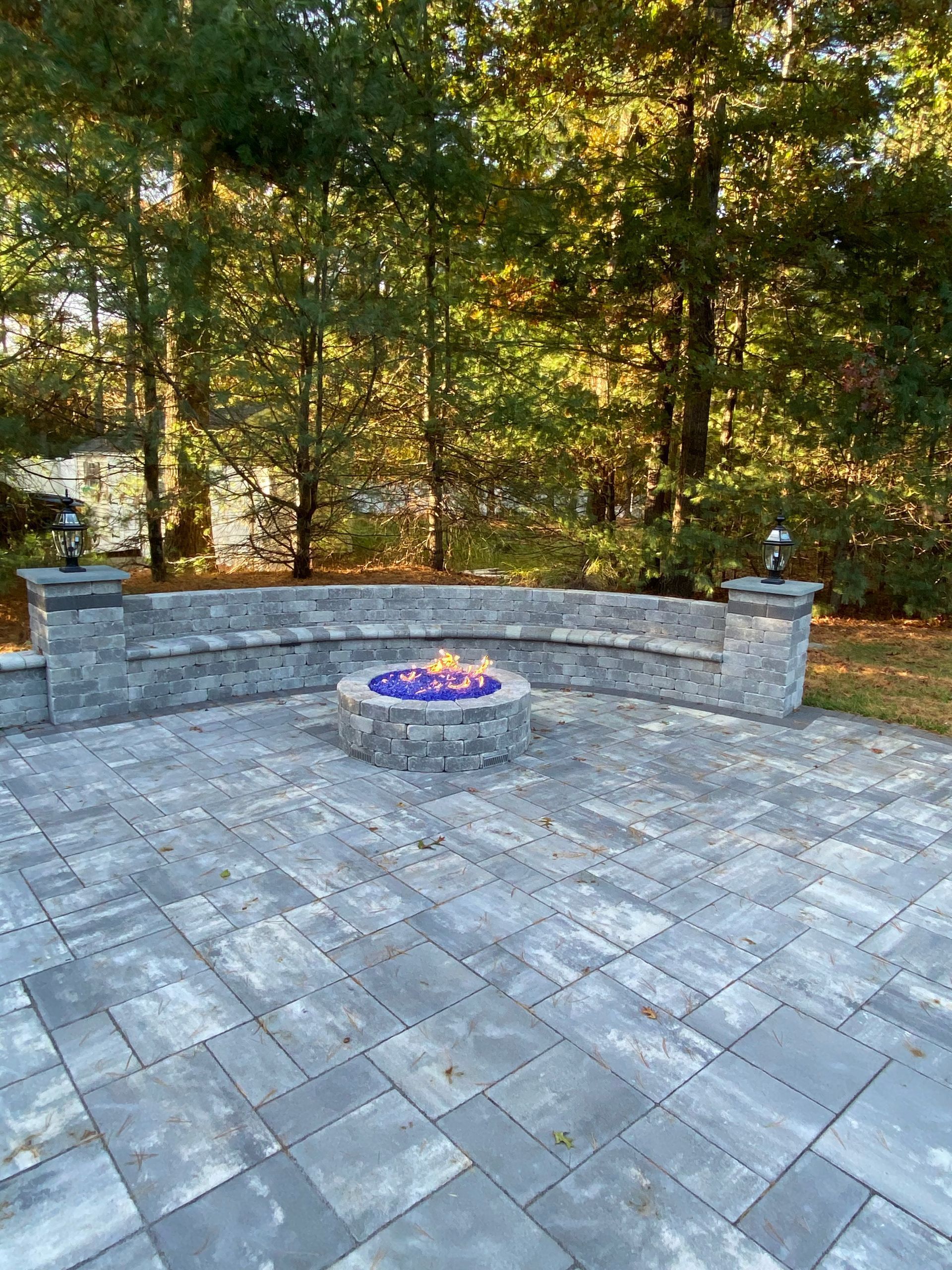 Stone patio with curved retaining wall and central fire pit, backed by autumn trees.
