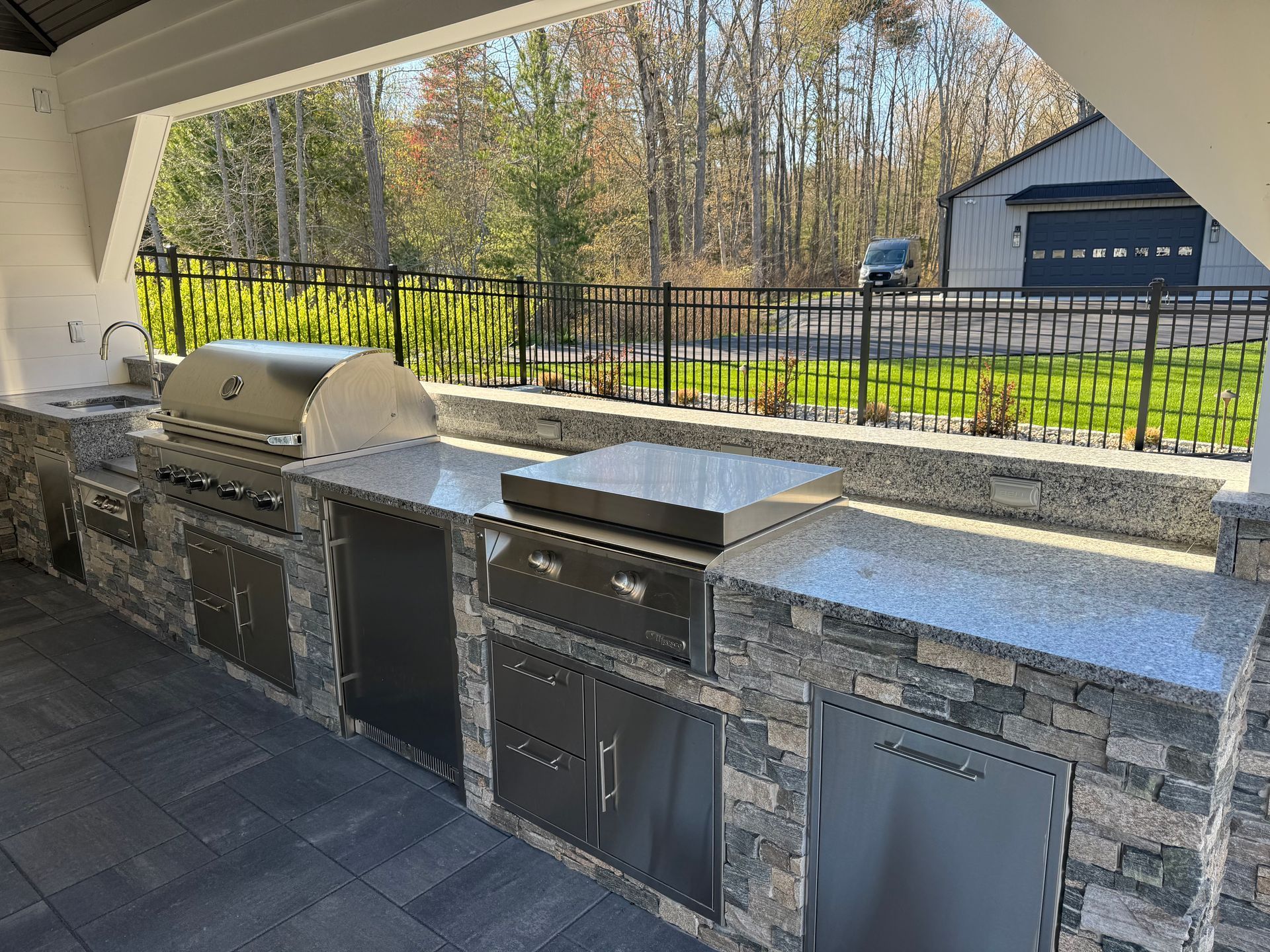 Outdoor stone patio kitchen with built-in grill, sink, cabinets, and black fence overlooking a yard and garage