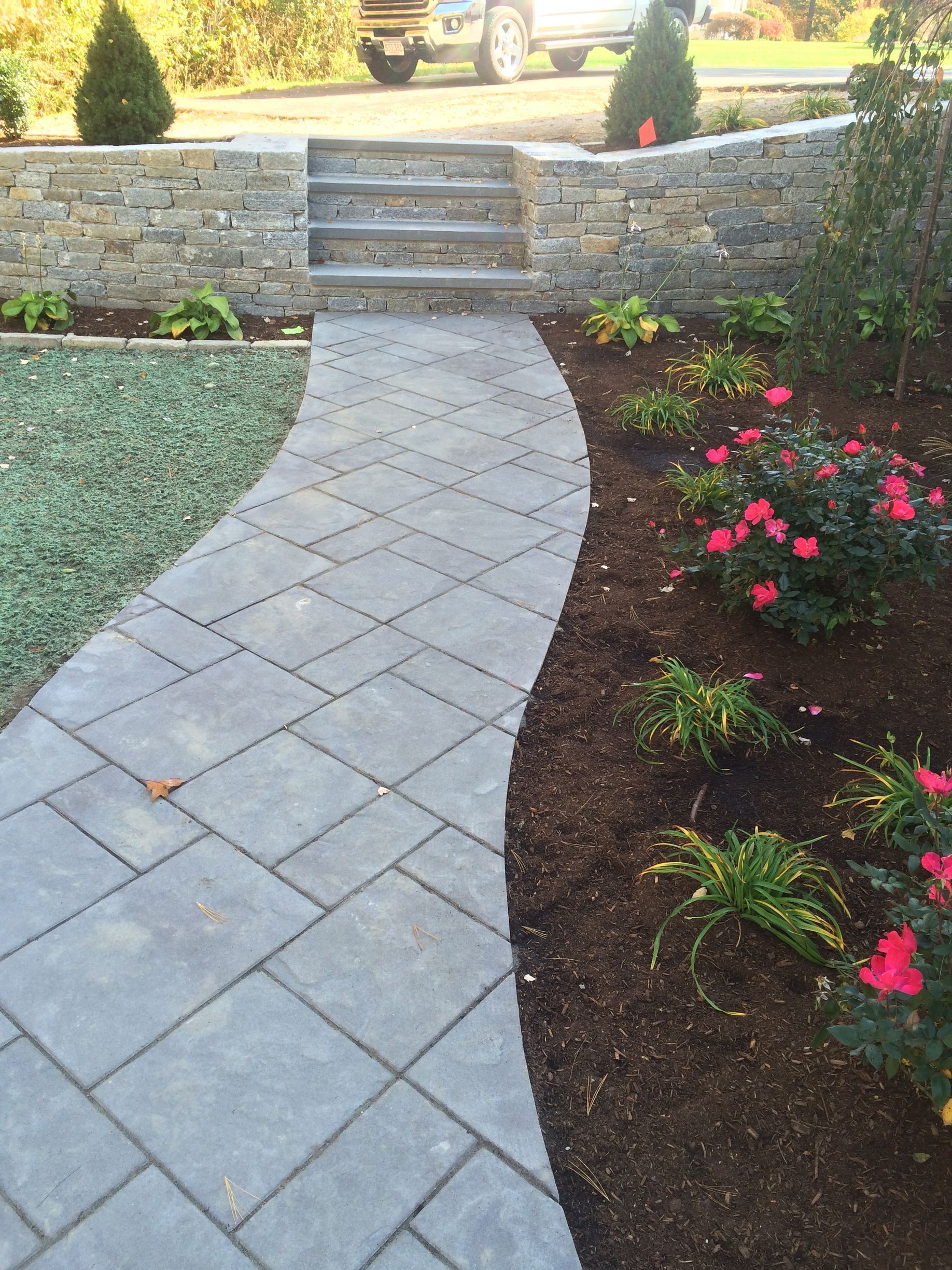Curved stone walkway leading to steps, bordered by mulch and pink flowers in a landscaped yard.