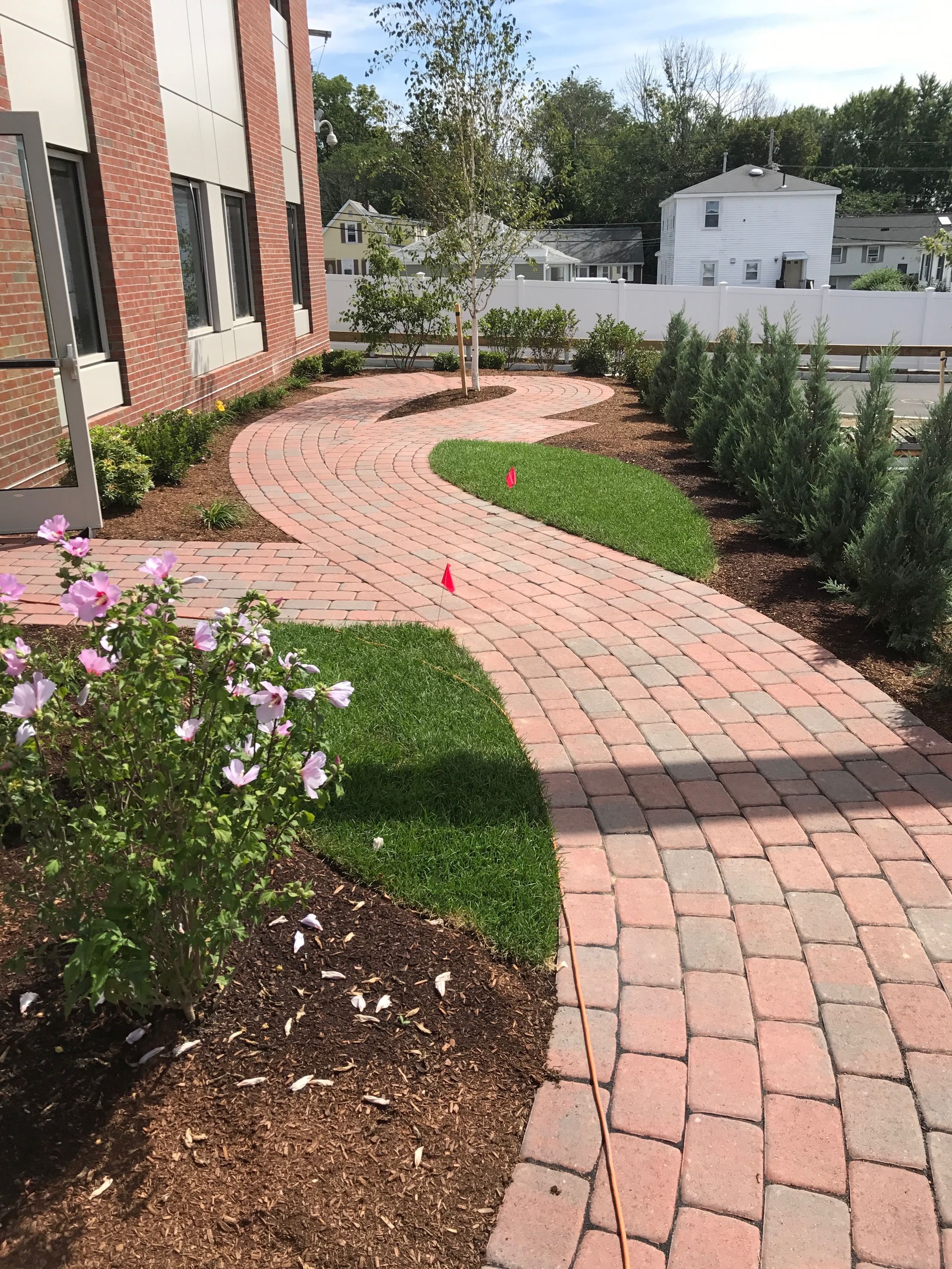 Curved brick walkway beside a building, with pink flowers, shrubs, and a landscaped garden border