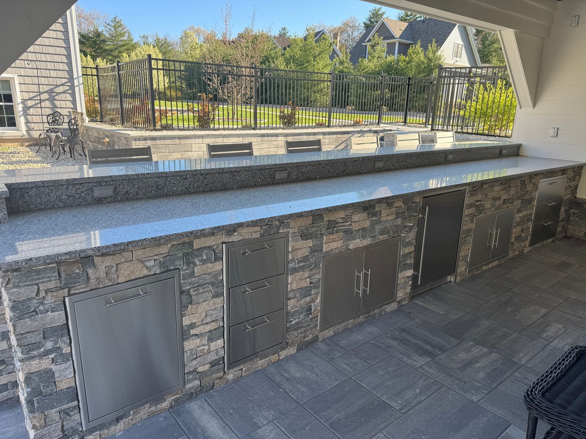 Stone outdoor bar with dark cabinets and a metal fence overlooking a landscaped yard
