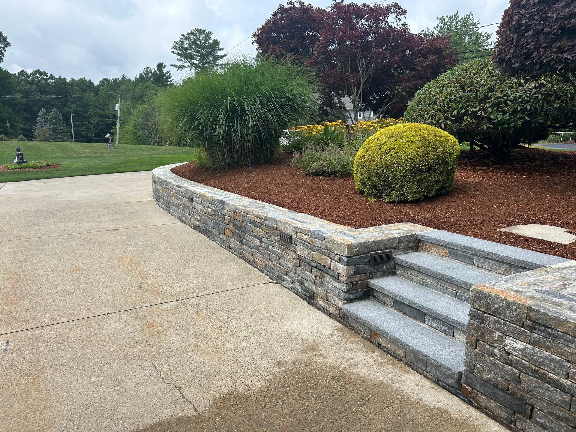 Landscaped garden with stone retaining wall, steps, shrubs, and a paved walkway.