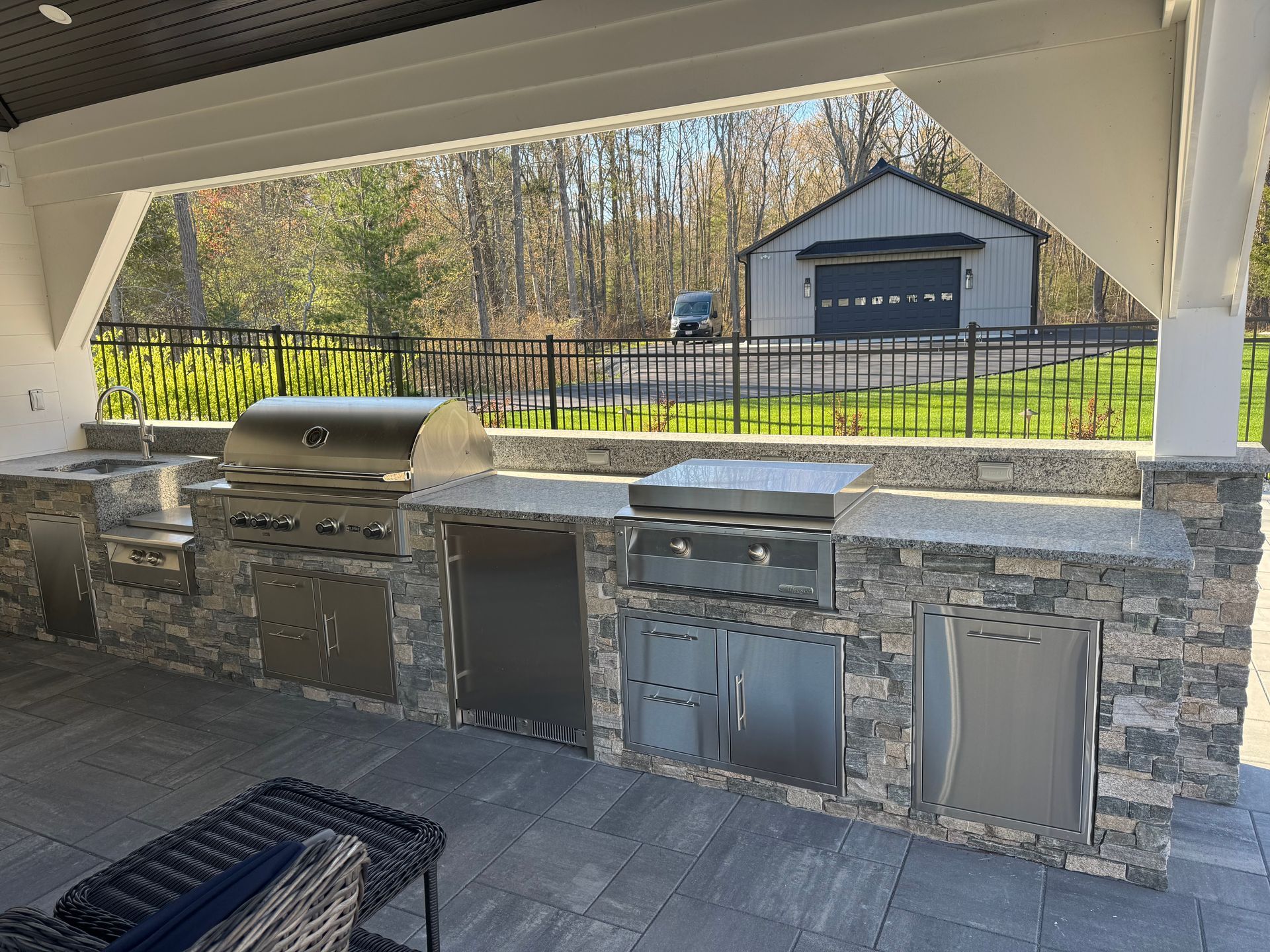 Covered outdoor kitchen with stone grill and cabinets under a pavilion, overlooking a fenced yard and shed