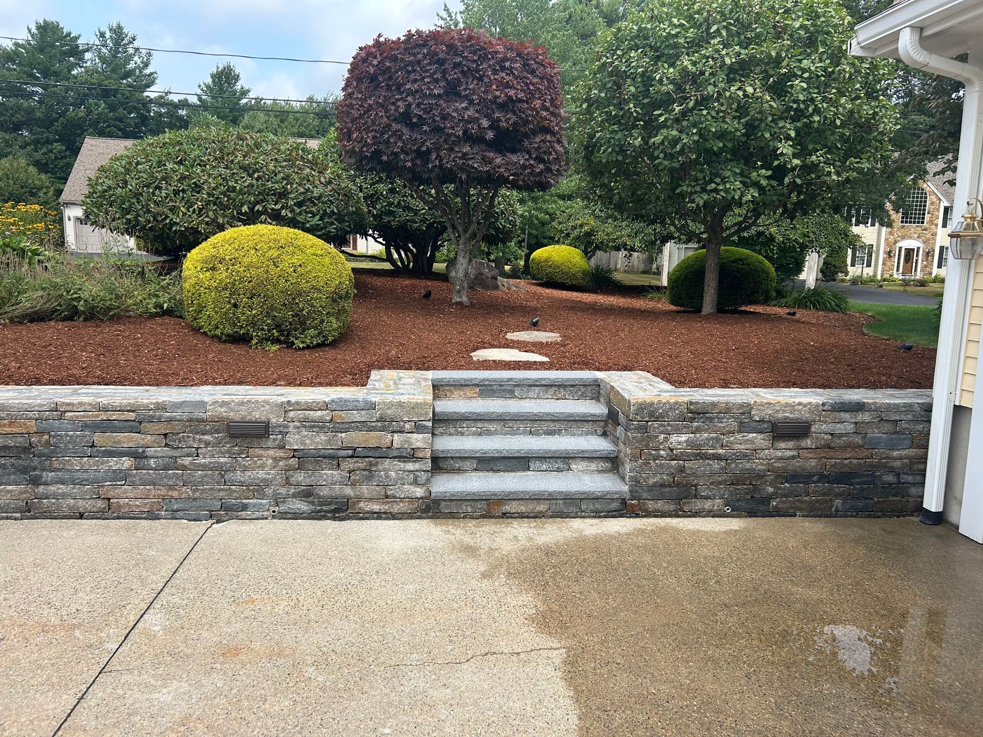 Front yard with layered stone retaining wall, mulch beds, trimmed shrubs, and a wet concrete driveway