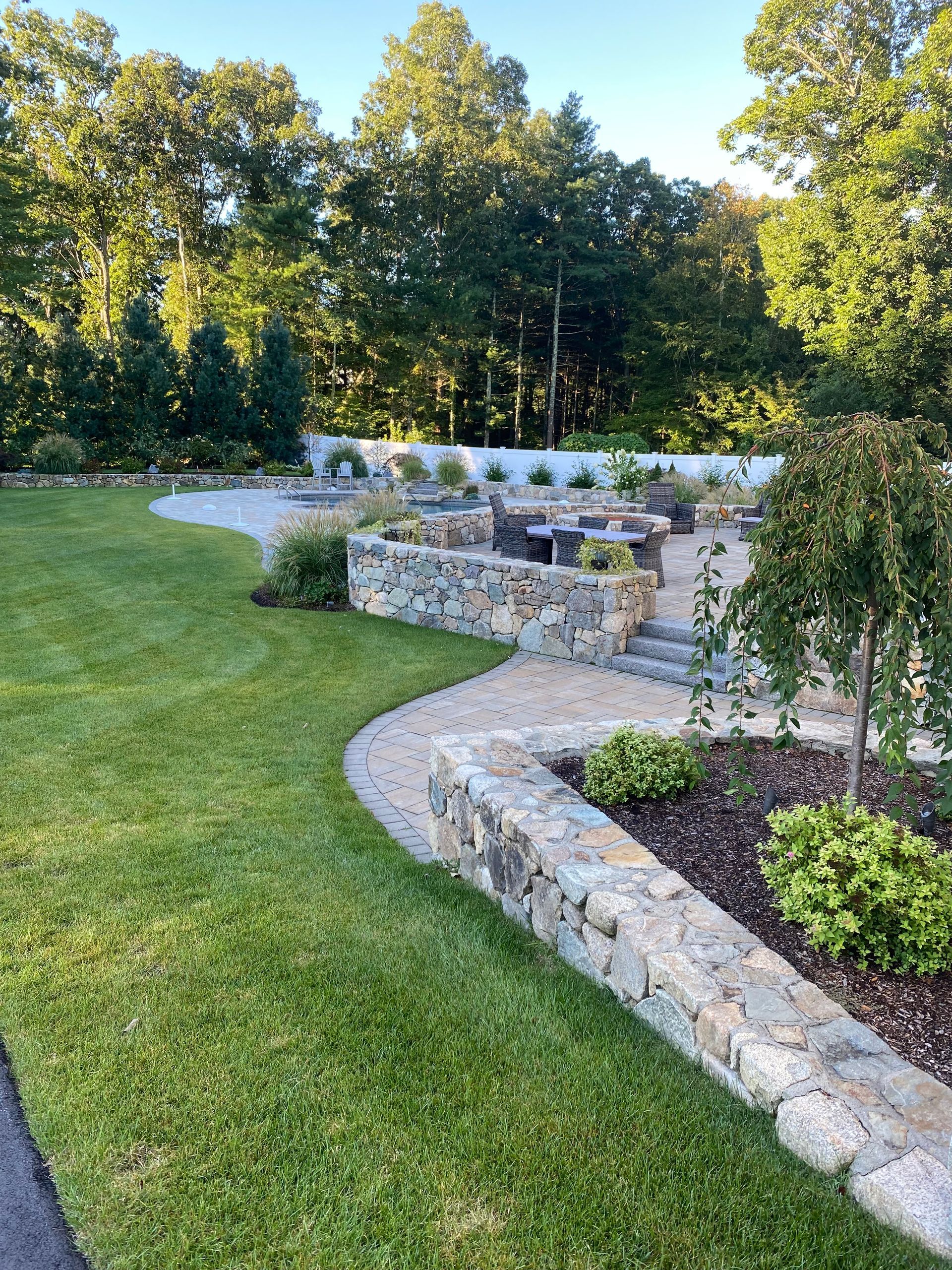 A lush green lawn with a stone walkway leading to a patio surrounded by trees.