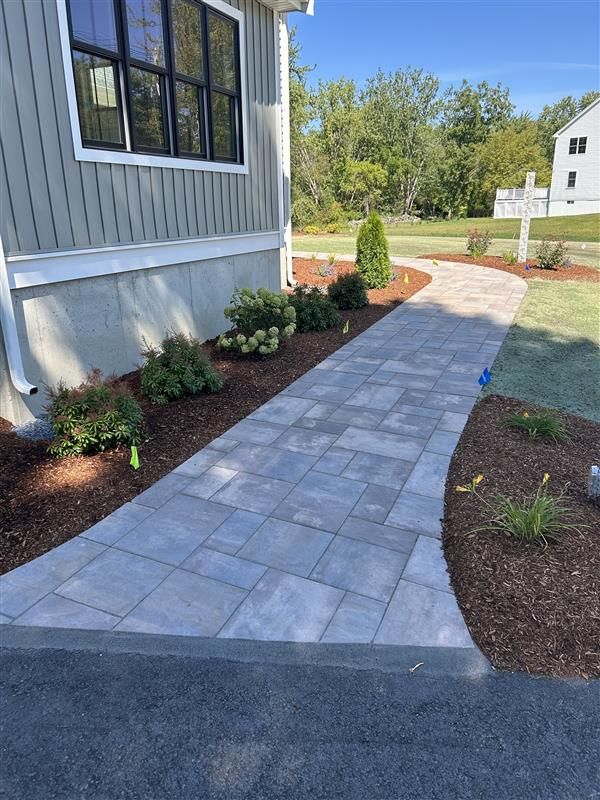 Gray paved walkway alongside a house with landscaping.