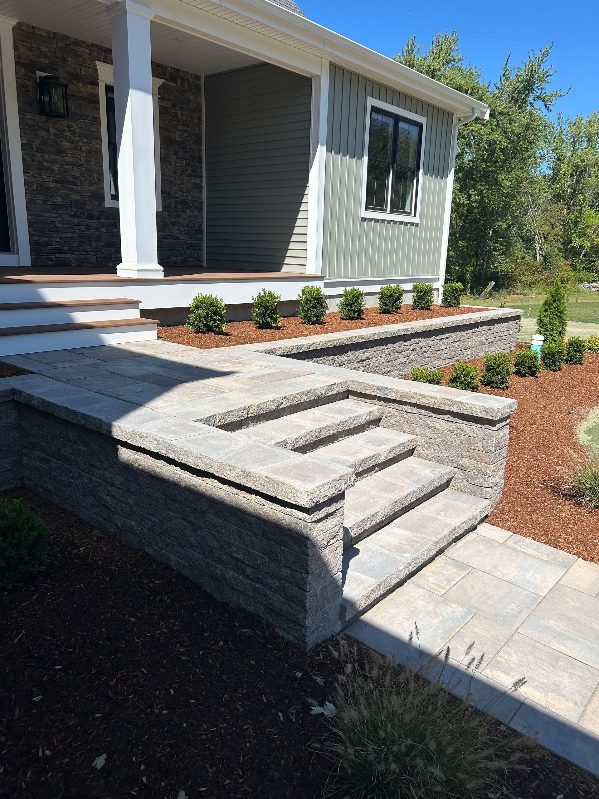 Stone steps lead to the porch of a house with gray siding and landscaping.