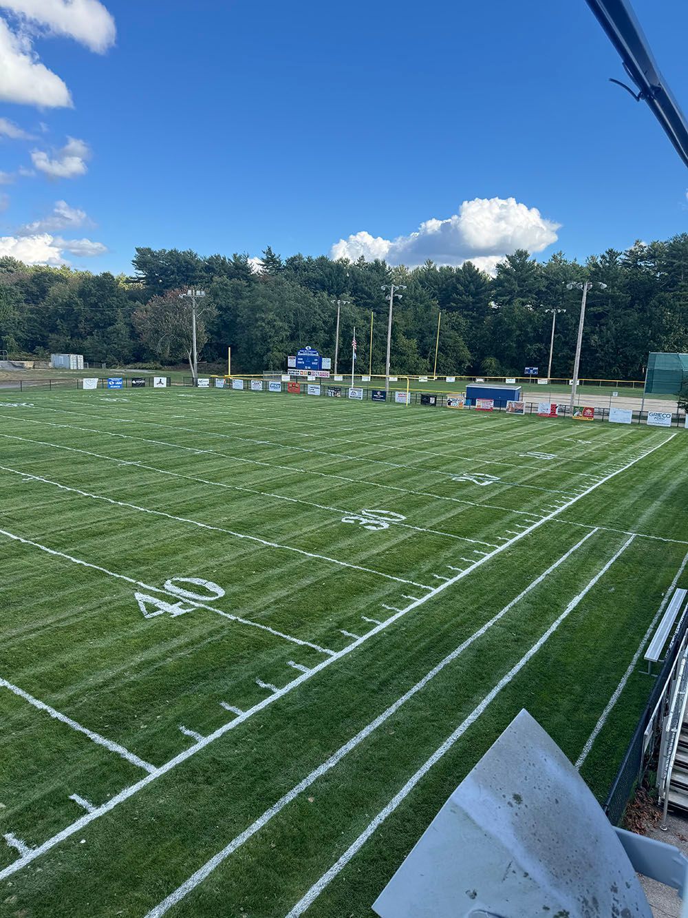 Football field with green grass, white yard lines, blue scoreboard, and trees under a blue sky