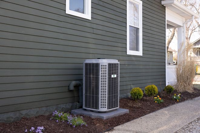 Outdoor HVAC unit beside a dark green house wall and window, next to a gravel path and shrubs