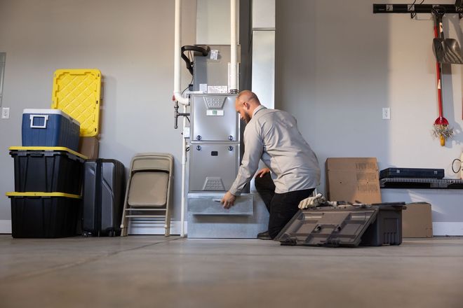 Man kneels beside a gray safe in a garage, handling its lower door amid stacked storage bins.