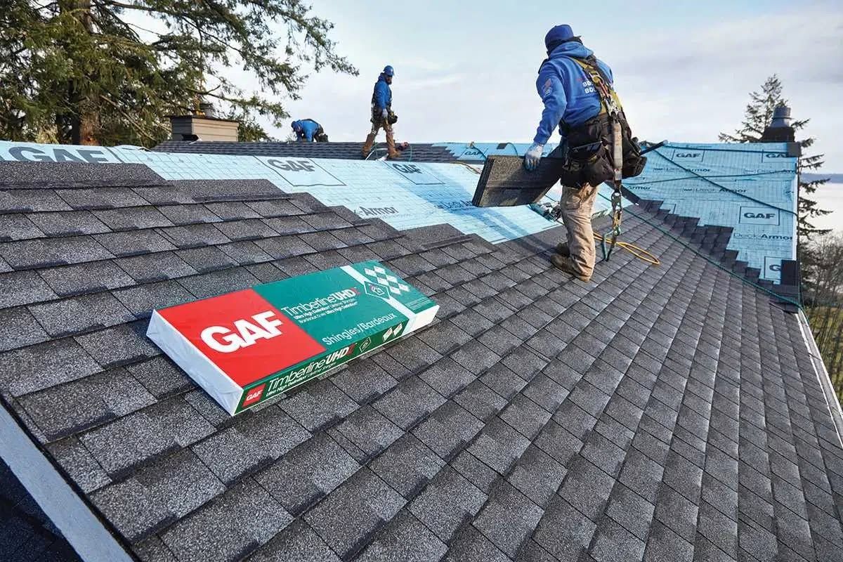 Workers installing shingles on a steep roof with a GAF roofing package in the foreground