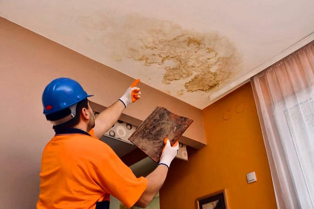 Worker in orange uniform and blue hard hat repairs a water-stained ceiling with a trowel.