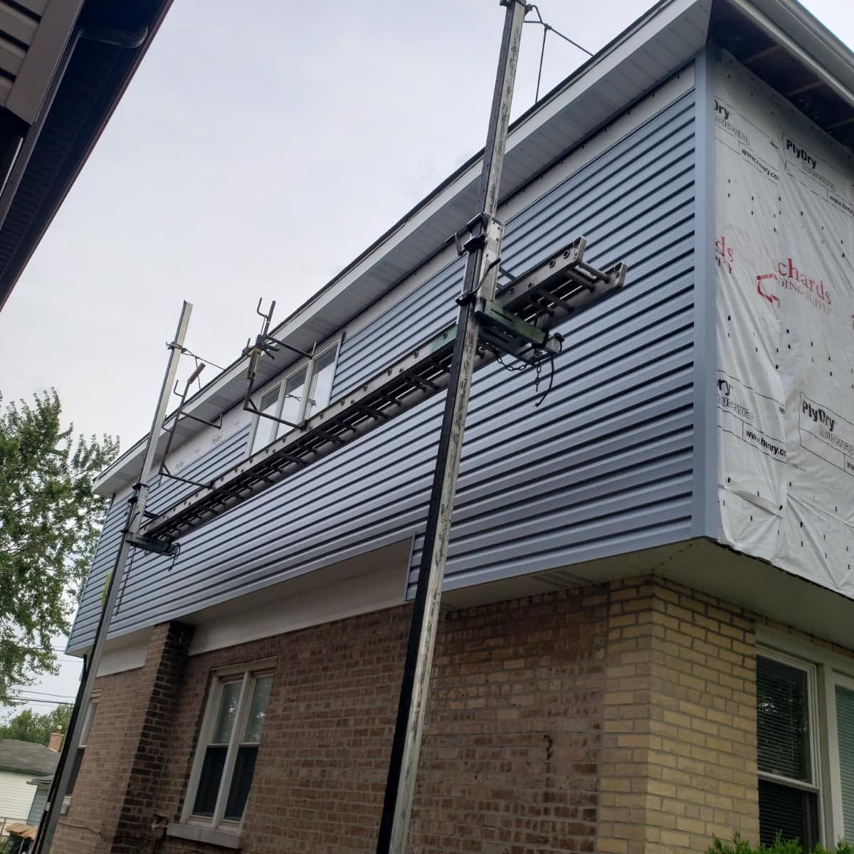Scaffold around a brick building with gray siding being installed on the upper wall