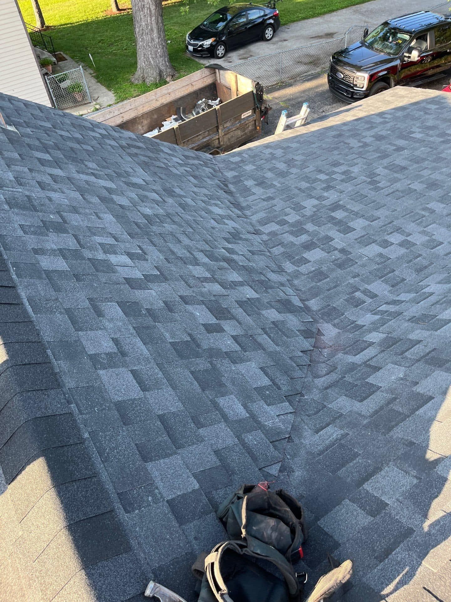 Rooftop view of gray shingles with a tool bag near the edge, overlooking a driveway and parked cars.
