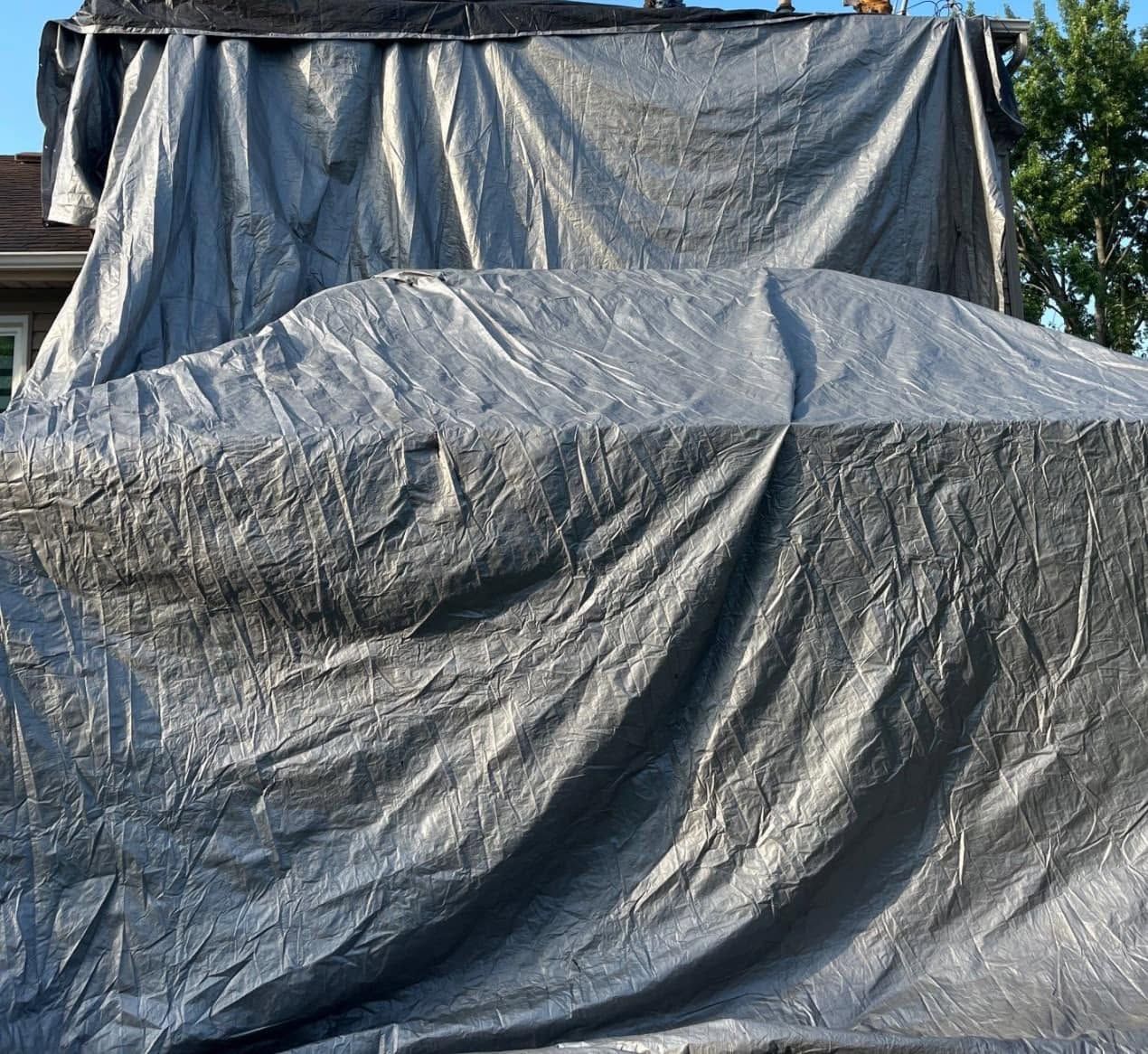 Large object covered with a gray tarp outdoors, with a larger dark tarp draped behind it.