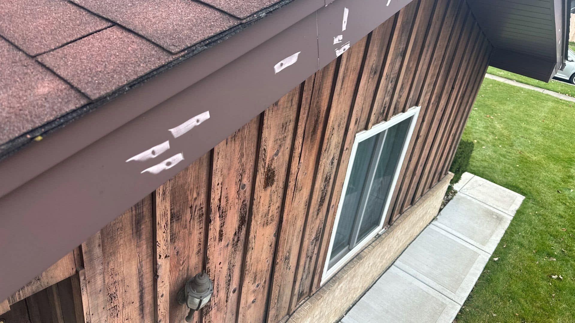 Exterior of a house with weathered wood siding, a window, roof edge, and a small concrete path by green lawn