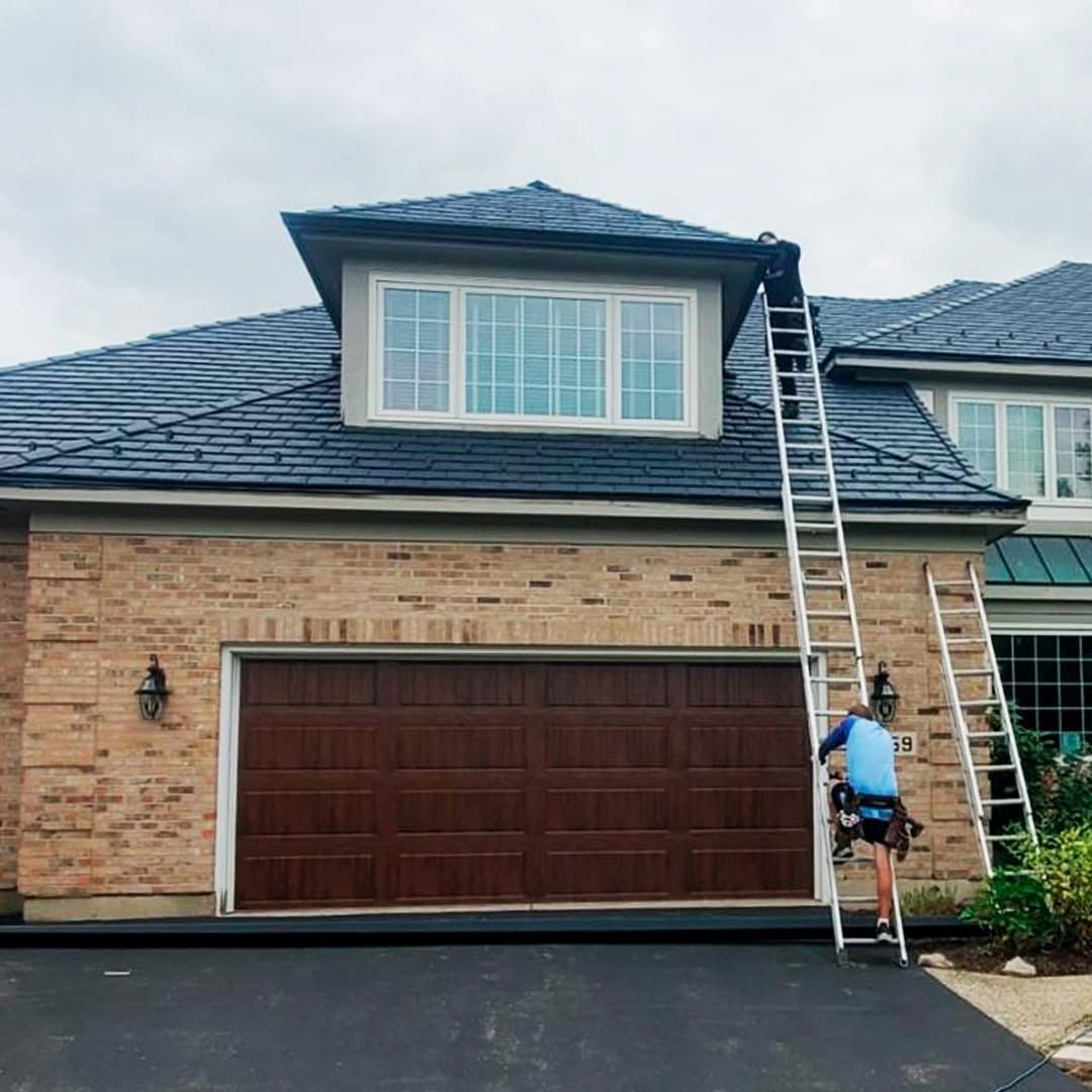 Worker on a ladder cleaning gutters on a brick house with a dark roof and garage door.