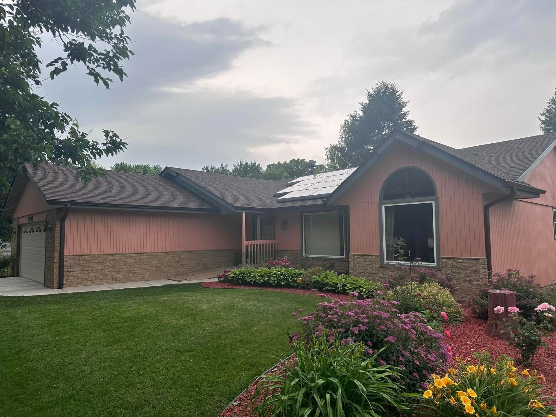 Single-story brick house with pink shutters, arched entry, and flower garden under a cloudy sky