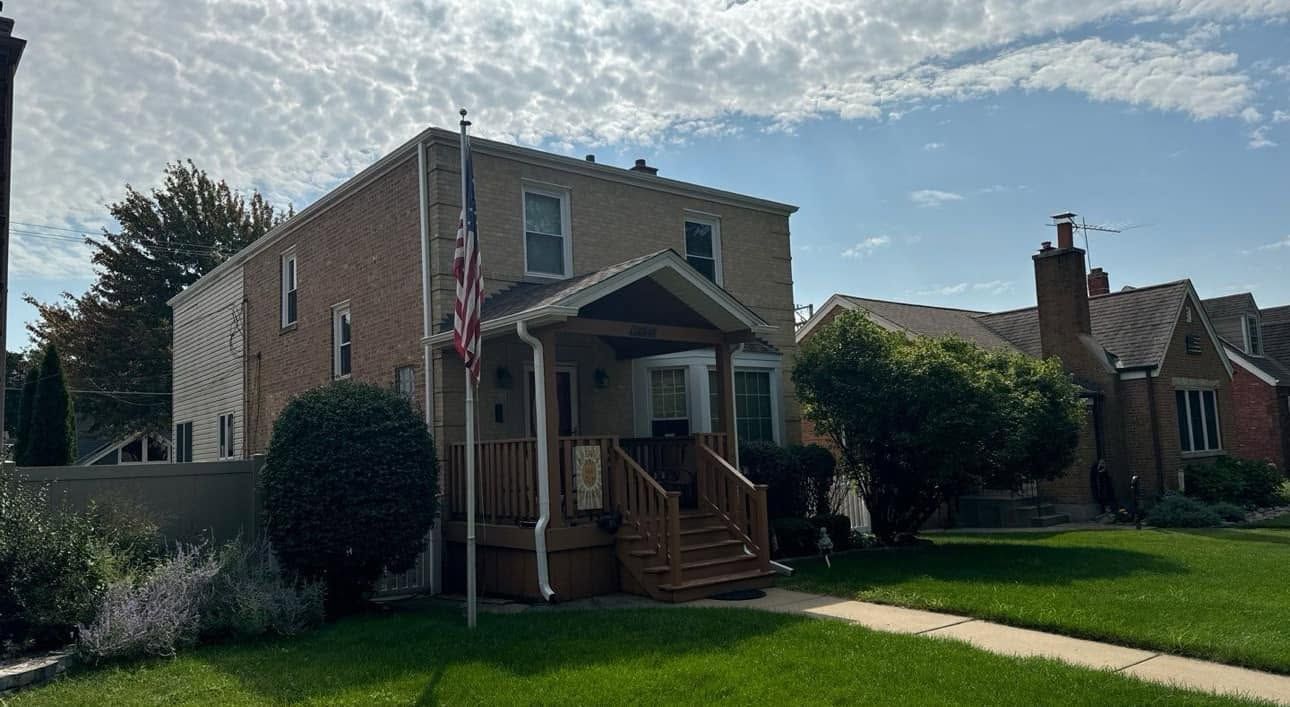 Brick two-story house with front porch, American flag, and green lawn under a cloudy sky