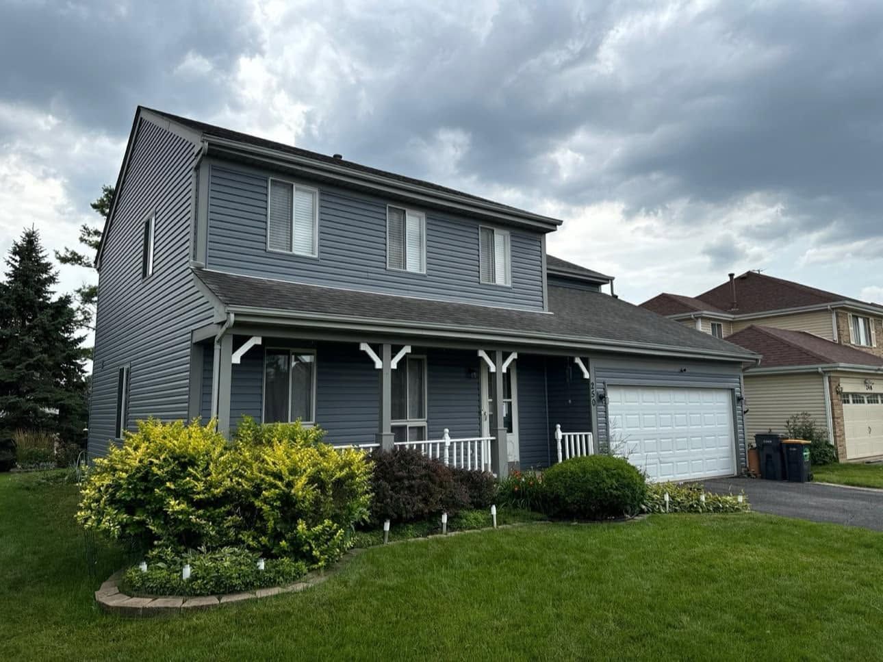 Two-story gray house with white garage, front porch, and cloudy sky above a green lawn