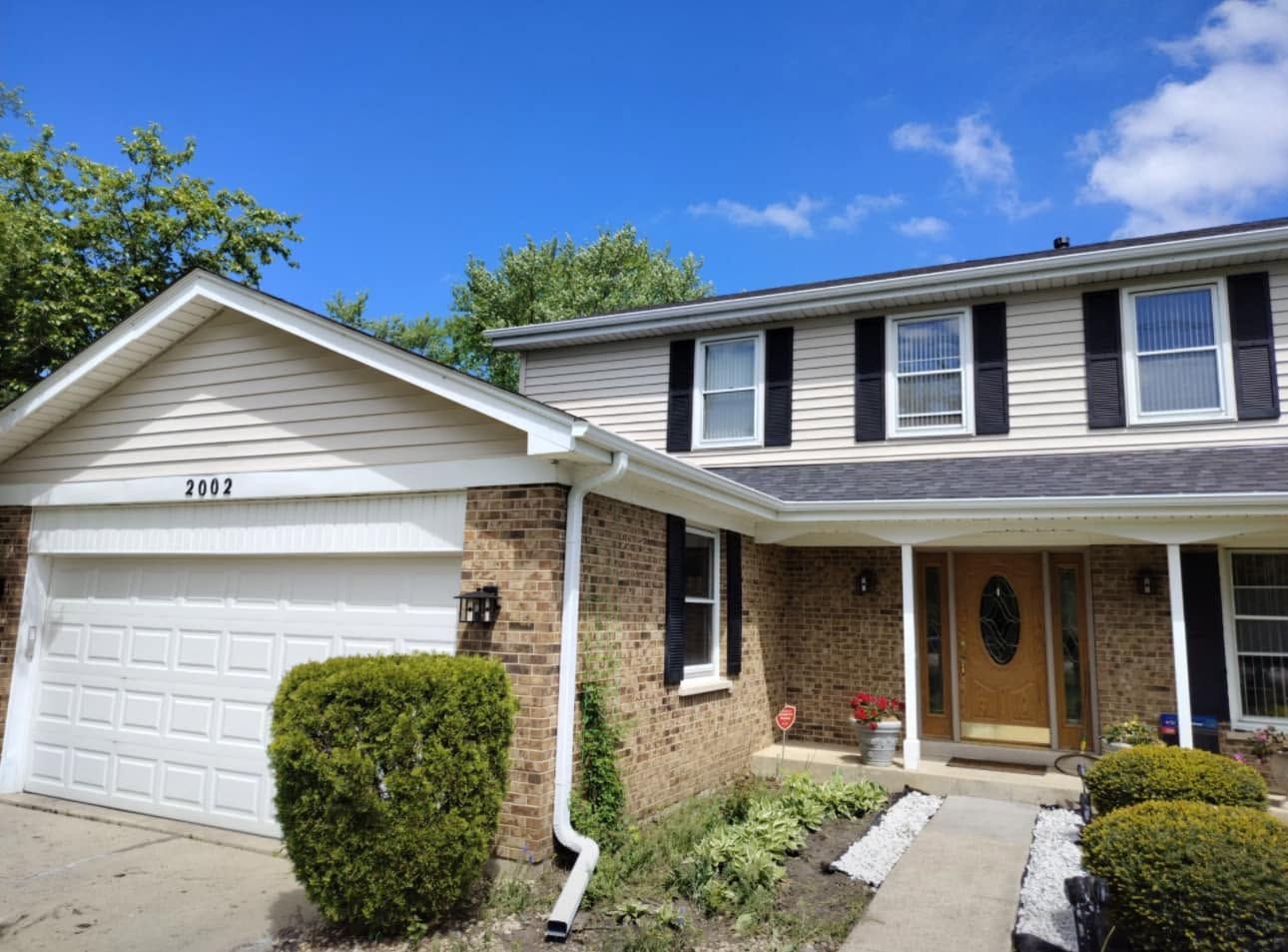 Suburban two-story brick house with white garage, front porch, and neatly trimmed bushes under a blue sky