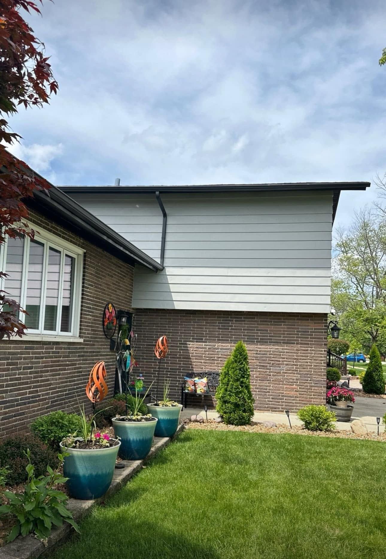 Brick-and-siding house exterior with potted plants and a green lawn under a cloudy sky