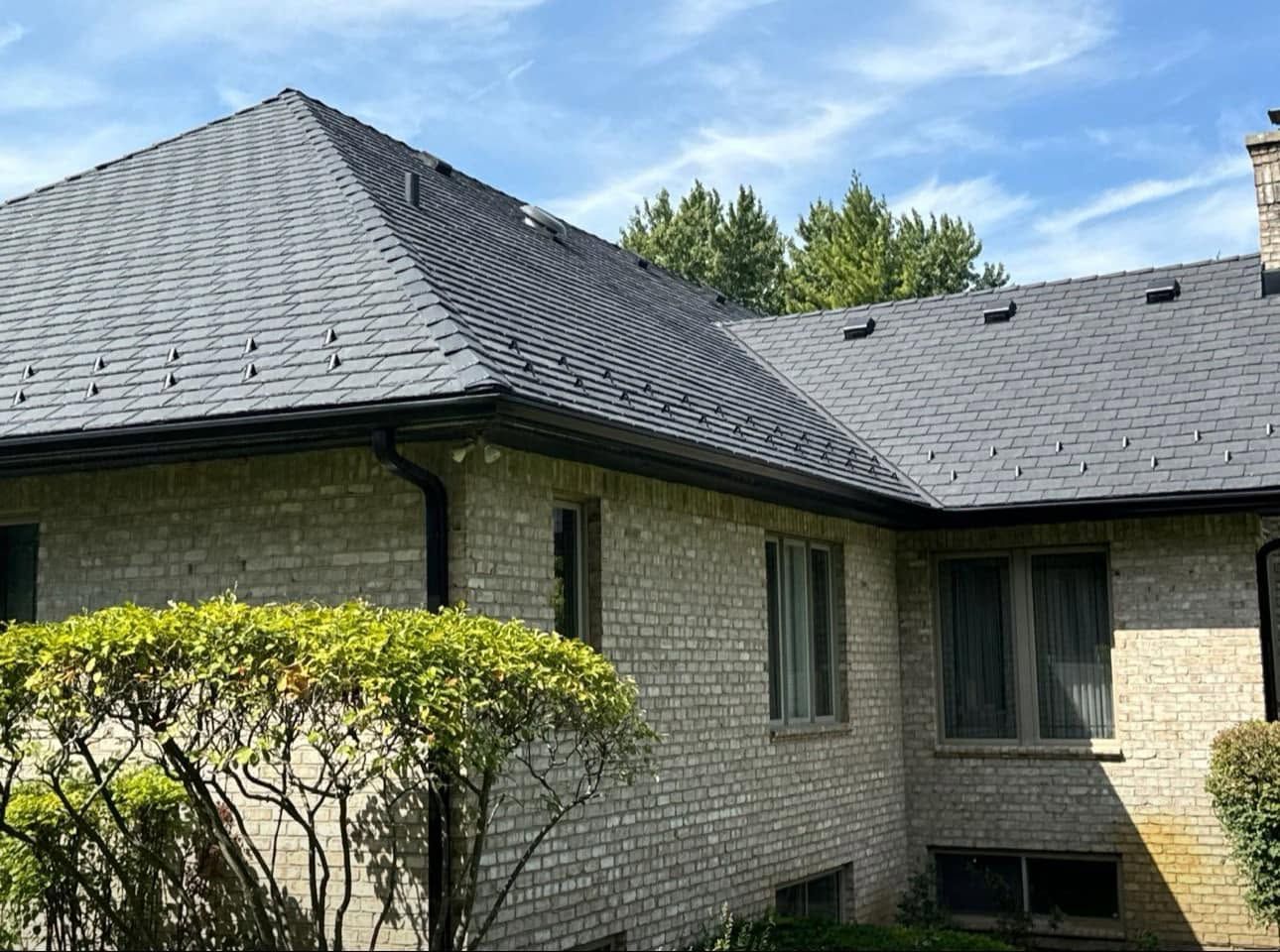 Brick house with dark gray roof, green shrub, and blue sky above.