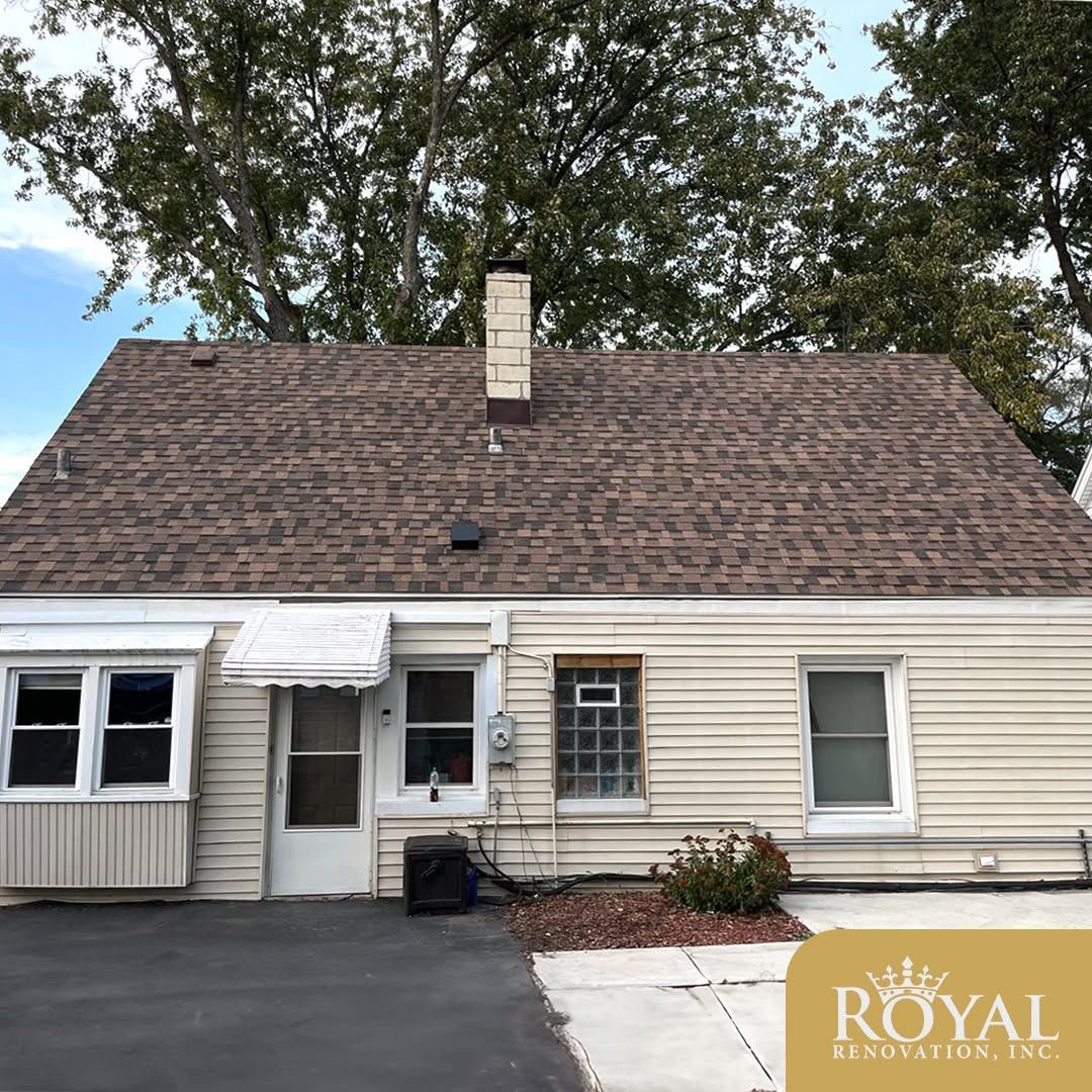 Small beige house with a brown shingle roof, front door, windows, and driveway under trees