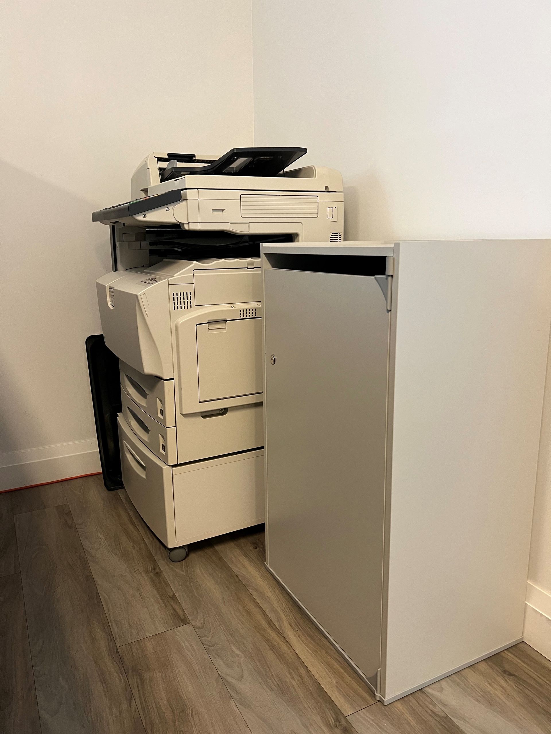 A printer is sitting in a corner of a room next to a cabinet.