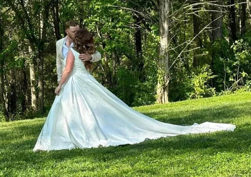 A bride and groom are kissing in the grass . the bride is wearing a long white dress.