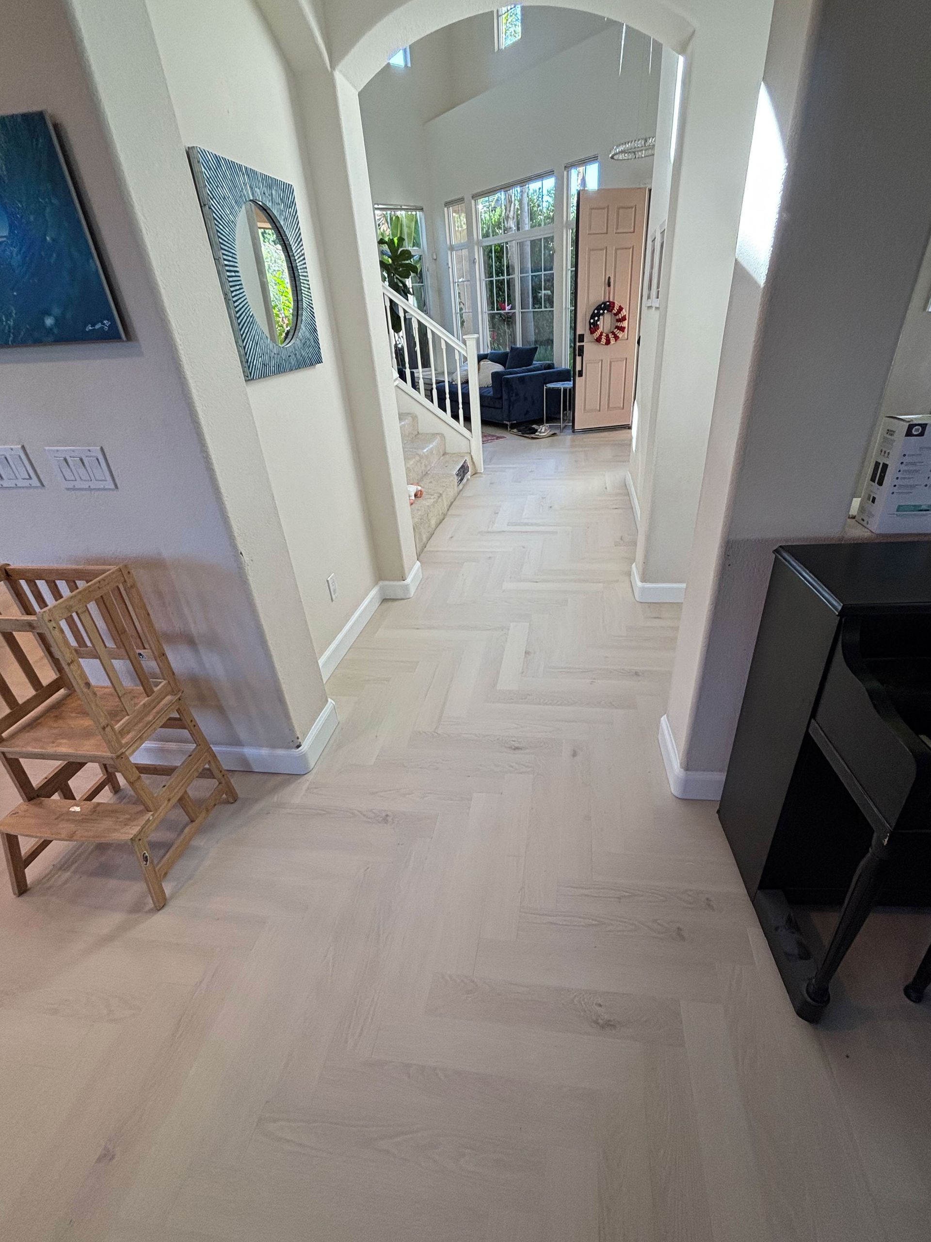 Bright hallway with white arches, light wood floor, and chairs along the left wall leading to a sunlit room.