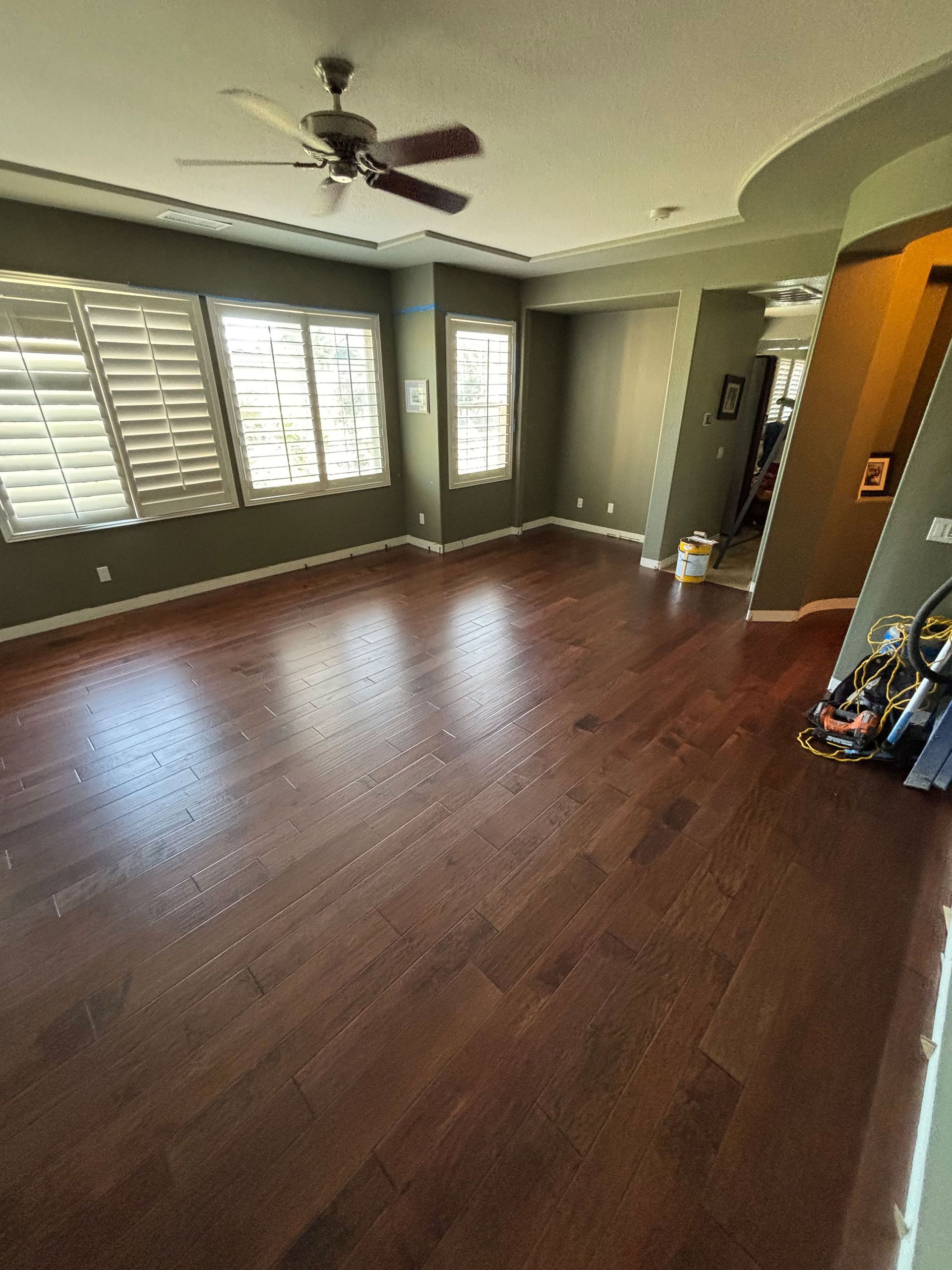 Empty living room with dark wood floors, gray walls, large windows, and ceiling fan.