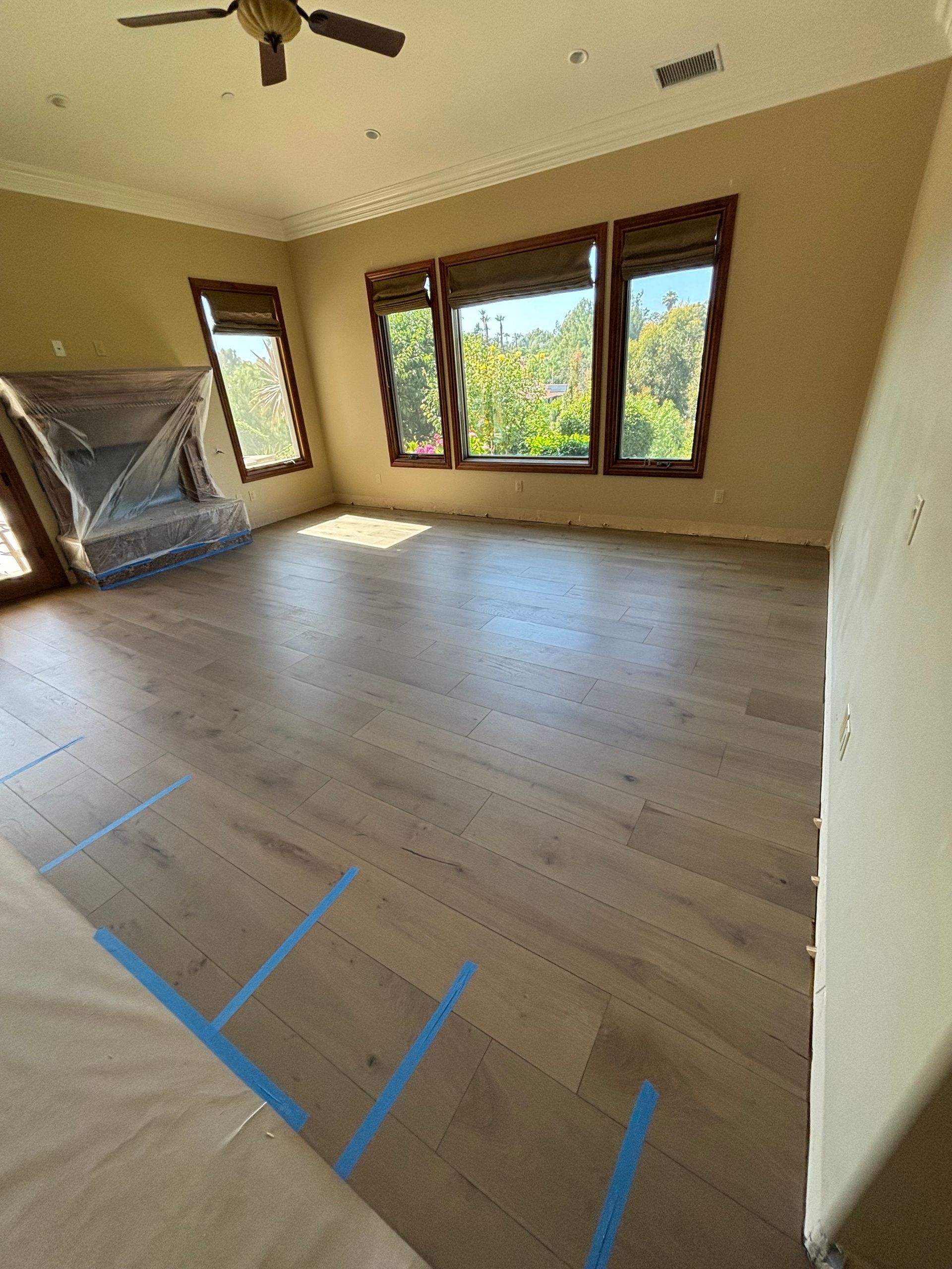 Empty beige bedroom with wood floor, ceiling fan, and large windows letting in daylight.