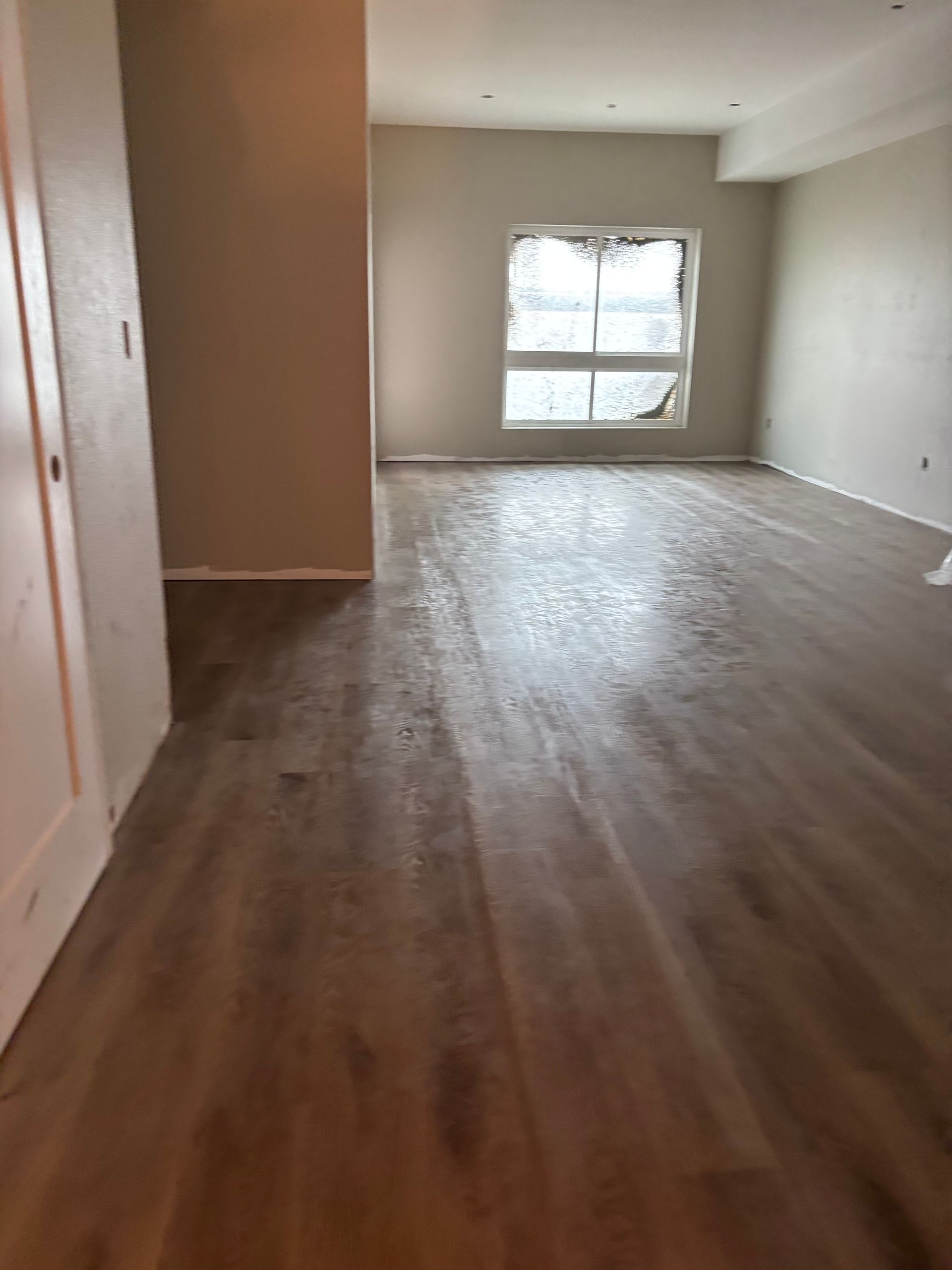 Empty room with wood flooring, beige walls, and a window with light curtains.