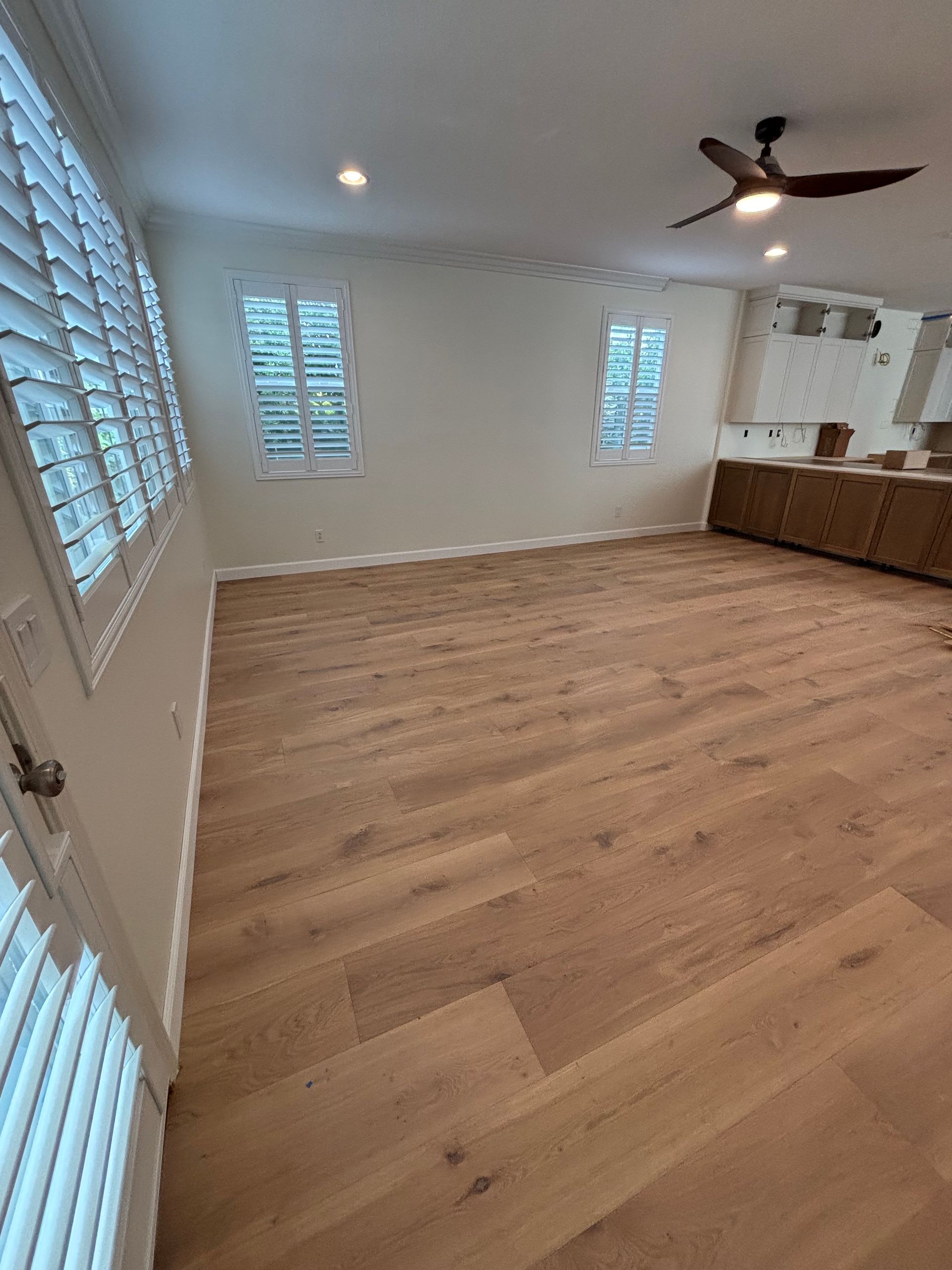 Empty living room with light wood floors, white walls, plantation shutters, ceiling fan, and kitchen area in back