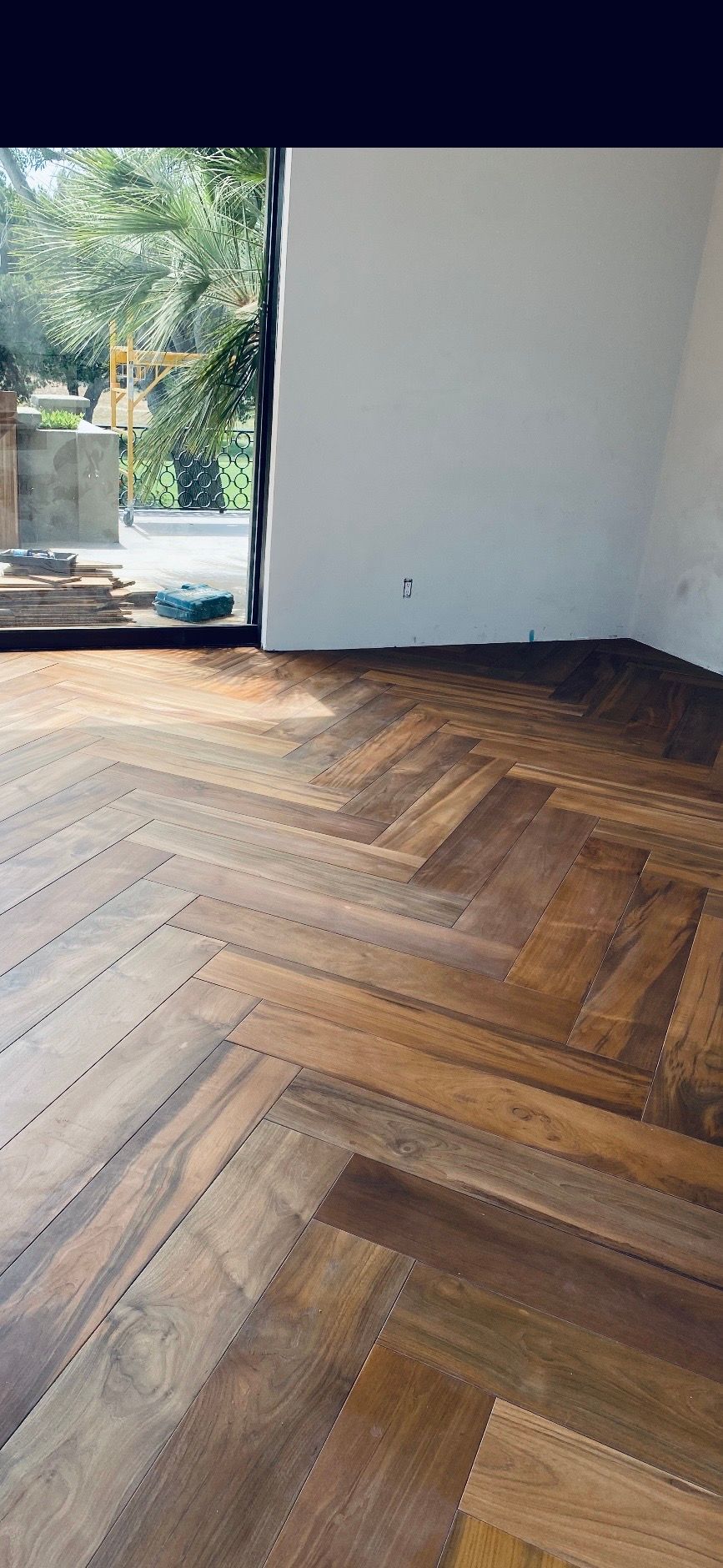 Empty room with dark herringbone wood floor, white walls, and a large window opening to greenery.