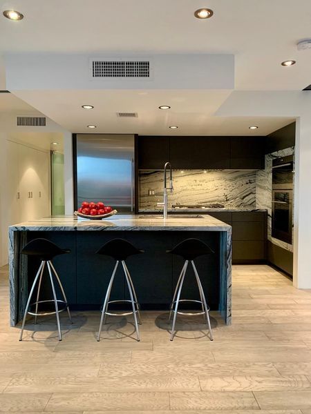 Modern kitchen with island, three black stools, stainless steel appliances, and marble countertops.