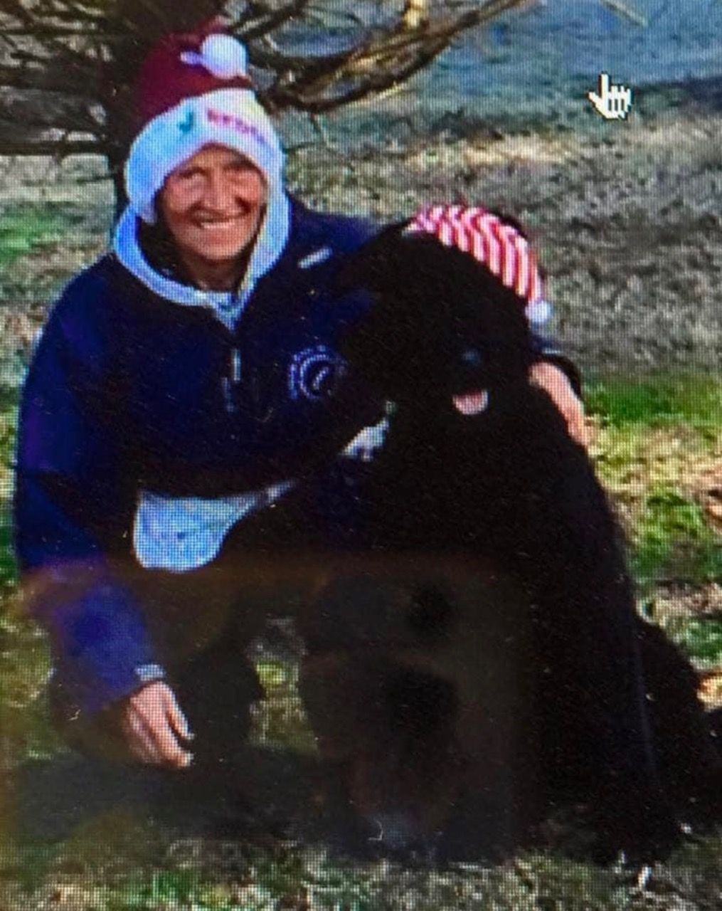 A man wearing a santa hat is kneeling down next to a black dog.