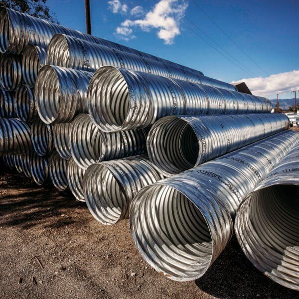 Piles of corrugated metal pipes, outdoors, stacked on a gravel surface, with a blue sky background.