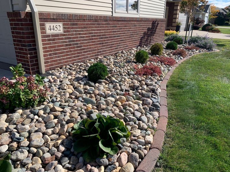 Landscaped bed with rocks and plants in front of a brick house, bordered by brick edging.
