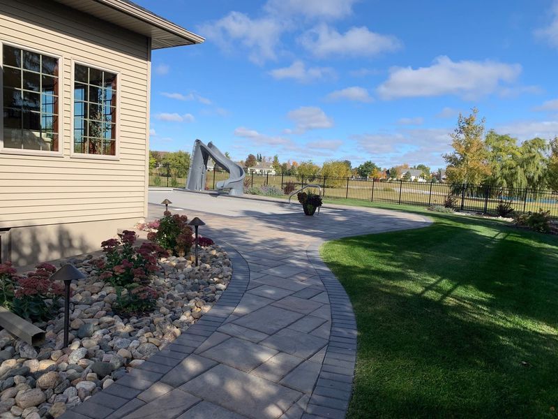 Beige house with a stone pathway leading into a sunny yard with sculptures and a fence.