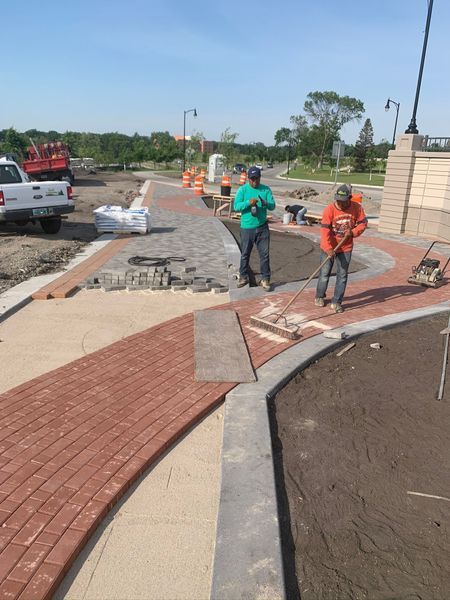 Workers laying brick pavers on a walkway, outdoors. One operates a tamper, the other stands.
