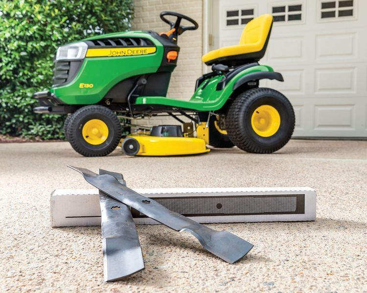Green John Deere riding mower with new blades on concrete near a garage.