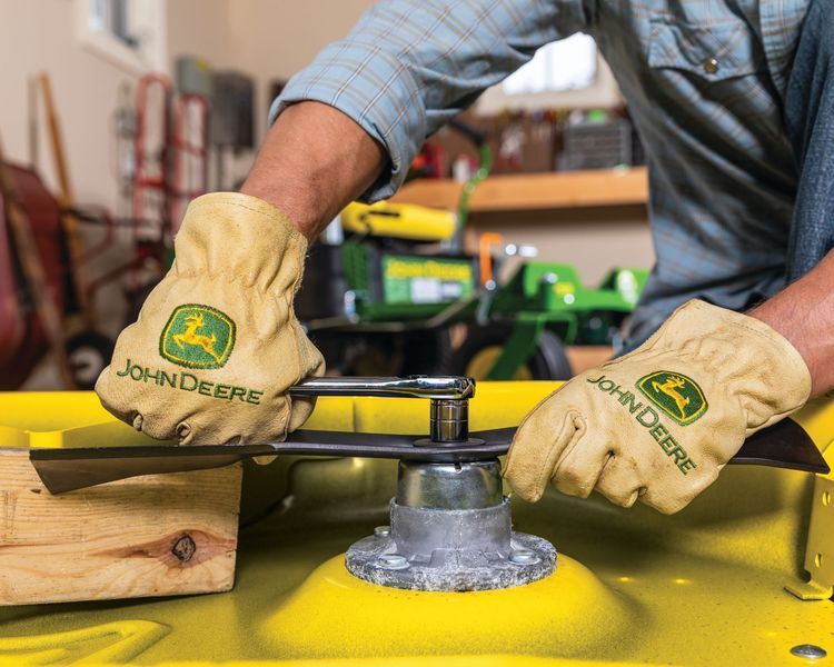 Person wearing John Deere gloves removing a lawn mower blade with a wrench in a garage.