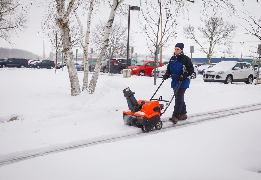 Person using an orange snowblower to clear a snowy path in a parking lot.