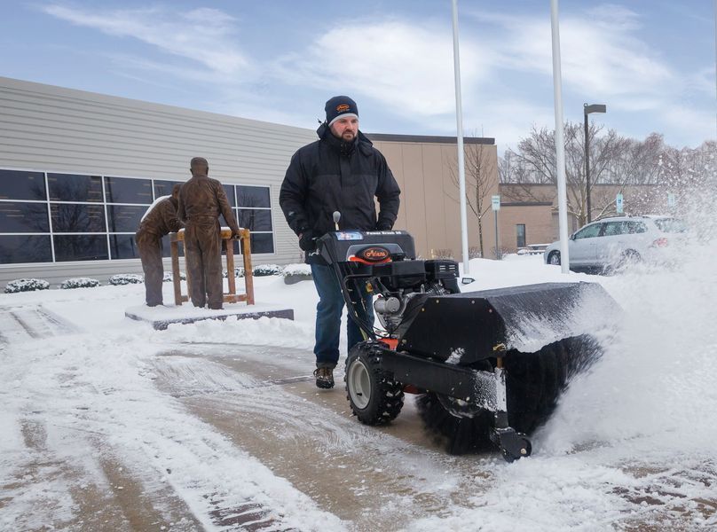A person using a snowblower, clearing snow from a walkway. Building and sculpture in the background.