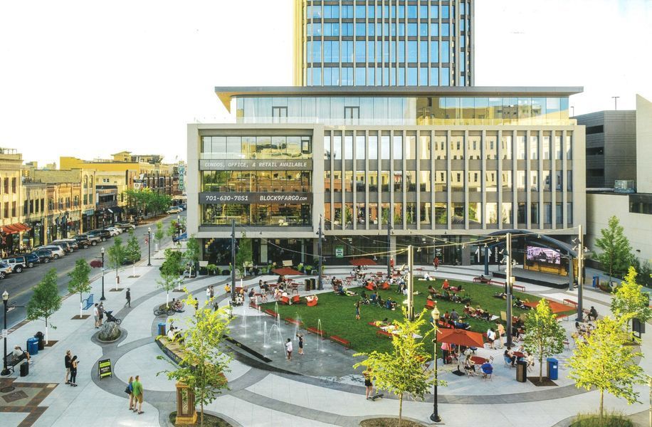 People gather in a plaza with a green space, water feature, and modern building.